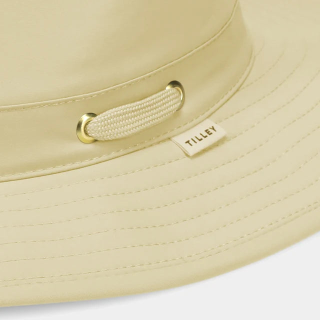 Close-up of a light beige wide-brim sun hat with a matching fabric chin strap and gold-tone metal hardware, featuring a small sewn-on 'TILLEY' brand tag.