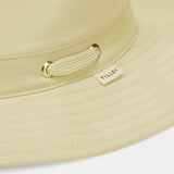 Close-up of a light beige wide-brim sun hat with a matching fabric chin strap and gold-tone metal hardware, featuring a small sewn-on 'TILLEY' brand tag.